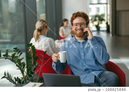 Smiling businessman talking phone with client while sitting in modern office and working on laptop Smiling businessman talking phone with client while sitting in modern office and working on laptop 108825898
