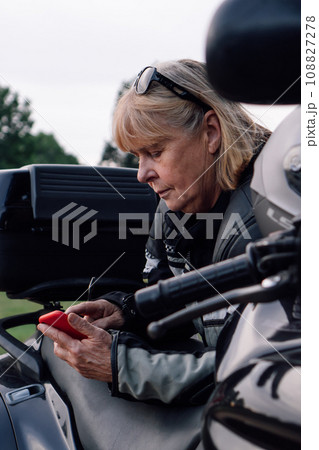 Communication in travel. Mature blonde on motorcycle, holding mobile phone in nature during stop. 108827278