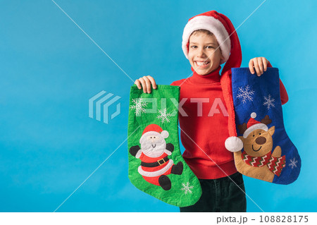 Child boy in red sweater and Santa hat with Christmas socks on blue background with space for text. Xmas and holidays concept mockup. Generation alpha and gen alpha children. Child boy in red sweater and Santa hat with Christmas socks on blue background with space for text. Xmas and holidays concept mockup. Generation alpha and gen alpha children. 108828175
