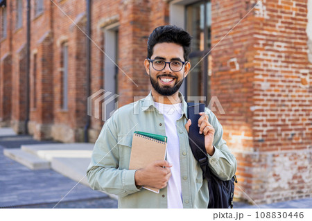 Portrait of young successful smiling student, Indian man close up smiling and looking at camera, man standing outside university campus in casual clothes with backpack and books in hands. 108830446