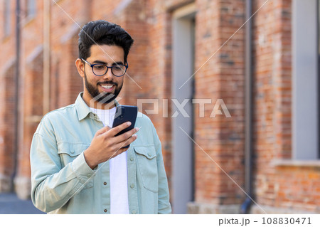 A young man walks through the city with a phone in his hands, a businessman in casual clothes uses an application on a smartphone, smiles, reads online pages, types a text message. A young man walks through the city with a phone in his hands, a businessman in casual clothes uses an application on a smartphone, smiles, reads online pages, types a text message. 108830471