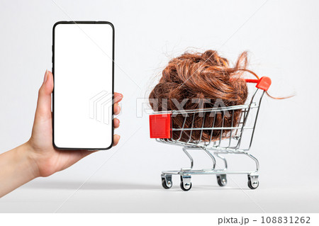 A female's hand holds a smartphone with a white screen. A shopping cart filled with brown hair falling down. White background. Mock up. The concept of buying and selling hair 108831262