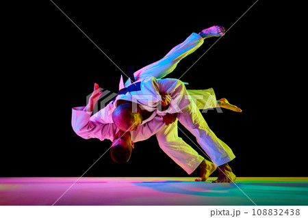 Portrait of two male sportsmen, martial arts fighters in white kimono fighting performing techniques in motion in neon light isolated black background. 108832438