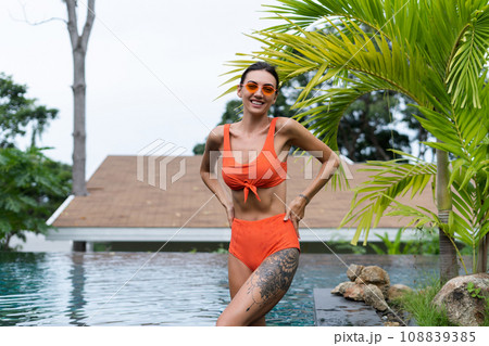 Beautiful european women in two pieces orange bikini swimsuit in perfect shape, good body, tanned, with trendy sunglasses outside at tropical swimming pool and palm leaf tree 108839385