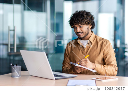 Man studying online remotely, businessman improving qualifications at the workplace inside office, writing down data in a notebook, watching a video course, satisfied with high results, smiling. Man studying online remotely, businessman improving qualifications at the workplace inside office, writing down data in a notebook, watching a video course, satisfied with high results, smiling. 108842057