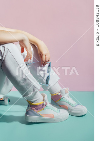 Close up female legs in white jeans and retro style high-top multicolor sport sneakers shoes sitting on the skateboard on turquoise and pink background. Vintage retro fashion style of 80s - 90s vibes. 108842139