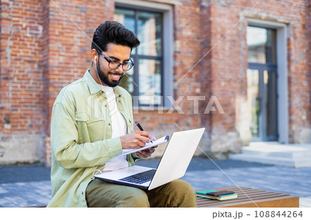 Young man studying remotely sitting on a bench outside a university building, a student in headphones watching a video online course, talking using a video call, writing down data in a notebook. Young man studying remotely sitting on a bench outside a university building, a student in headphones watching a video online course, talking using a video call, writing down data in a notebook. 108844264