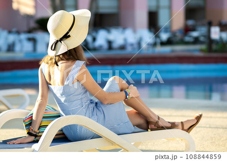 Young woman wearing light summer dress and yellow straw hat sitting outside near hotel swimming pool on summer sunny day. Young woman wearing light summer dress and yellow straw hat sitting outside near hotel swimming pool on summer sunny day. 108844859