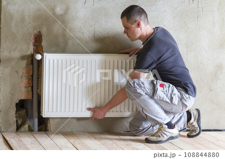 Young handsome professional plumber worker installing heating radiator in an empty room of a newly built apartment or house. Construction, maintenance and repair concept. Young handsome professional plumber worker installing heating radiator in an empty room of a newly built apartment or house. Construction, maintenance and repair concept. 108844880