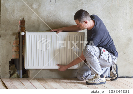 Young handsome professional plumber worker installing heating radiator in an empty room of a newly built apartment or house. Construction, maintenance and repair concept. Young handsome professional plumber worker installing heating radiator in an empty room of a newly built apartment or house. Construction, maintenance and repair concept. 108844881