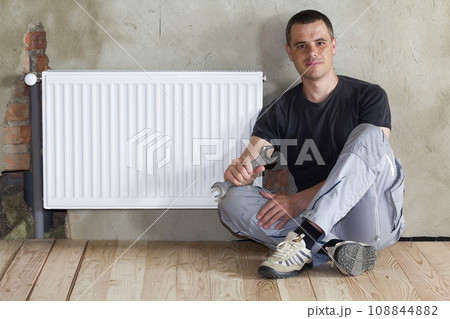 Young handsome plumber sitting on floor with wrench in hand near successfully installed heating radiator in empty room of a newly built apartment or house. Construction, maintenance and repair concept 108844882
