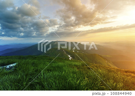 Wide summer mountain view at sunrise. Glowing orange sun raising in blue cloudy sky over green grassy hill soft grass and distant mountain range covered with morning mist. Beauty of nature concept. 108844940