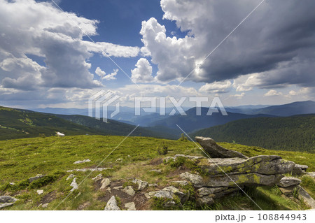 Wide panorama of lit by sun mountain plateau with green grass, patches of snow and big boulders on distant mountains under cloudy sky background. Beauty of nature, tourism and traveling concept. 108844943