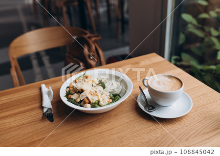 A white bowl full of Cezar salad with croutons, chicken and parmesan cheese. Cezar salad and cup of coffee on white plate over wooden table A white bowl full of Cezar salad with croutons, chicken and parmesan cheese. Cezar salad and cup of coffee on white plate over wooden table 108845042