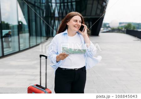 Beautiful redhead woman holding paper map and talking on smartphone outdoors. Lady with suitcase choosing route via paper map, navigating her urban journey 108845043
