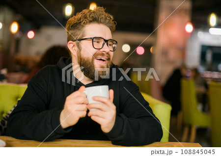 Bearded curly man smiling confident drinking coffee in restaurant or coffee shop. Millennial generation and Gen Y 108845045