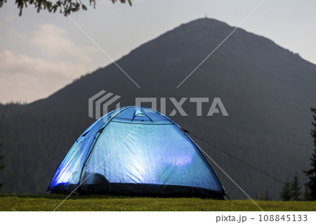 Tourist hikers bright blue tent on green grassy forest clearing among tall pine trees under clear morning sky on distant foggy woody mountain background. Tourism, summer camping in mountains at dawn. 108845133