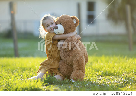 Pretty child girl tenderly embracing her teddy bear friend outdoors on green grass lawn. Concept of friendship values Pretty child girl tenderly embracing her teddy bear friend outdoors on green grass lawn. Concept of friendship values 108845514