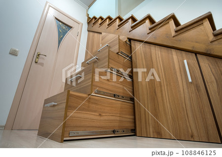Modern architecture interior with luxury hallway with glossy wooden stairs in multi-storey house. Custom built pullout cabinets on glides in slots under stairs. Use of space for storage. Modern architecture interior with luxury hallway with glossy wooden stairs in multi-storey house. Custom built pullout cabinets on glides in slots under stairs. Use of space for storage. 108846125