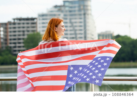 Happy young red haired woman with USA national flag on her shoulders standing outdoors in american city. Positive girl celebrating United States independence day. Happy young red haired woman with USA national flag on her shoulders standing outdoors in american city. Positive girl celebrating United States independence day. 108846364