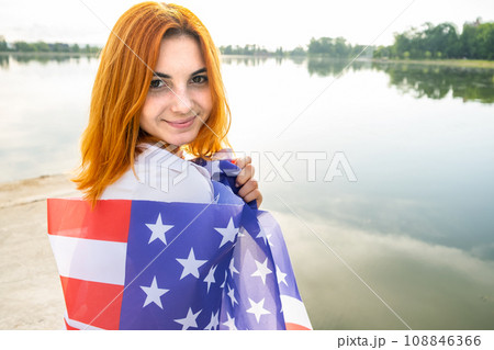 Happy smiling red haired girl with USA national flag on her shoulders. Positive young woman celebrating United States independence day. Happy smiling red haired girl with USA national flag on her shoulders. Positive young woman celebrating United States independence day. 108846366