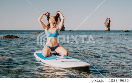 Woman sea sup. Close up portrait of happy young caucasian woman with long hair looking at camera and smiling. Cute woman portrait in a blue bikini posing on sup board in the sea 108849626
