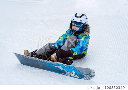 little boy sitting on snow putting his feet in snowboard bindings adjusting straps.  108850349