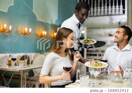 African american waiter bringing ordered dishes to couple at restaurant 108852412