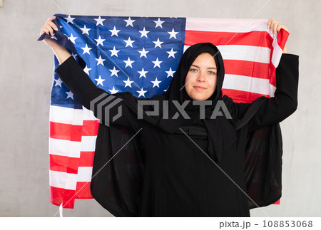 Balanced smiling Muslim woman in traditional black hijab holds flag of United States of America 108853068