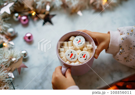 Unrecognizable little child girl holding cup with hot chocolate with marshmallows as snowman. Kid sitting near Christmas decorated window with lights. 108856011