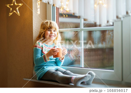 Little preschool girl holding cup with hot chocolate with marhsmallows. Happy child drinking sweet cocoa by window with Christmas lights in winter. Cozy family celebration of xmas. Little preschool girl holding cup with hot chocolate with marhsmallows. Happy child drinking sweet cocoa by window with Christmas lights in winter. Cozy family celebration of xmas. 108856017