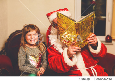 Little toddler girl talking to Santa Claus called Nikolaus or Weihnachtsmann in German. Happy smiling kid waiting for gifts. Santa with golden book. Xmas, childhood, holiday 108856248