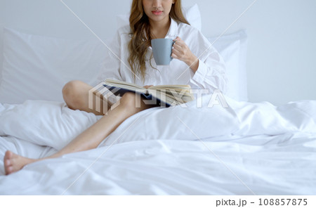 Young woman drinking coffee while reading a book sitting on the bed in the morning. 108857875