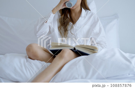 Young woman drinking coffee while reading a book sitting on the bed in the morning. Young woman drinking coffee while reading a book sitting on the bed in the morning. 108857876