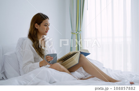 Young woman drinking coffee while reading a book sitting on the bed in the morning. Young woman drinking coffee while reading a book sitting on the bed in the morning. 108857882