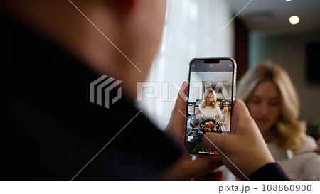 A man takes a picture of his beautiful blonde with a mobile phone. The concept of romantic relationships. Comfortable lifestyle. A man takes a picture of his girlfriend with a mobile phone in a cafe A man takes a picture of his beautiful blonde with a mobile phone. The concept of romantic relationships. Comfortable lifestyle. A man takes a picture of his girlfriend with a mobile phone in a cafe 108860900