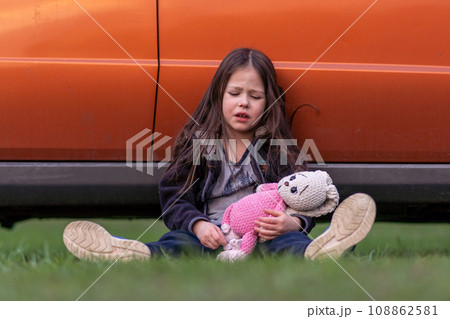 Children's problems. Little lonely girl is very upset, tired, holding toy in her hands, sitting on grass against car . Children's problems. Little lonely girl is very upset, tired, holding toy in her hands, sitting on grass against car . 108862581