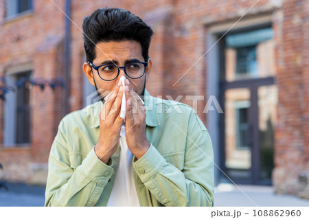 A man sneezes while sitting on a bench outside an office building, a young businessman has a runny nose and allergies, sick. A man sneezes while sitting on a bench outside an office building, a young businessman has a runny nose and allergies, sick. 108862960