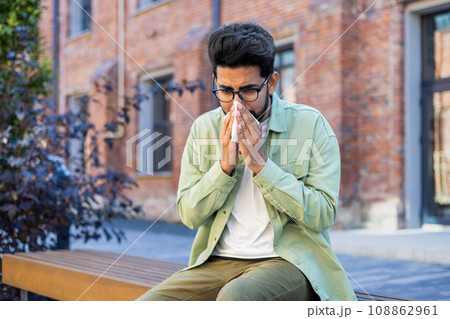 A man sneezes while sitting on a bench outside an office building, a young businessman has a runny nose and allergies, sick. A man sneezes while sitting on a bench outside an office building, a young businessman has a runny nose and allergies, sick. 108862961