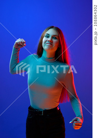 Young attractive woman standing looking at camera arguing and showing, clenching fist against blue background in neon light filter. 108864302