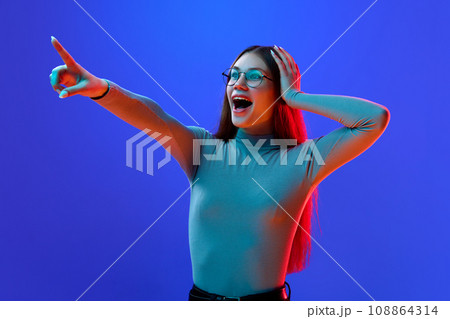 Wow. Surprised, delightful young girl, teenager in style glasses pointing that new sales season beginning isolated blue background in neon light. Concept of fashion. 108864314