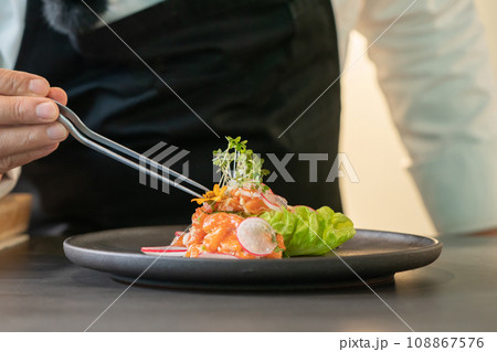 Tweezers chef in Chef's hand decorates Spicy Salmon Salad by micro herb on the food. 108867576