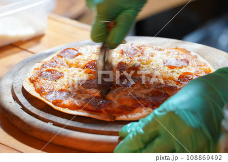 A person wearing green gloves is cutting thin-crust, small-size pizzas on a wooden tray with a pizza cutter. 108869492