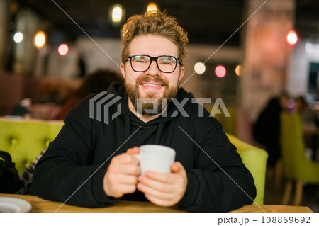 Bearded curly man smiling confident drinking coffee in restaurant or coffee shop. Millennial generation and Gen Y 108869692