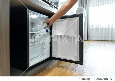 Man Hand opened mini small refrigerator and reached drinking plastic water bottle under the wood counter in the hotel resort bedroom. Man Hand opened mini small refrigerator and reached drinking plastic water bottle under the wood counter in the hotel resort bedroom. 108870015