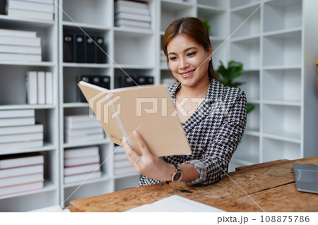 Portrait of a young Asian Woman showing a smiling face as she uses his notebook, computer and financial documents on her desk in the early morning hours. Portrait of a young Asian Woman showing a smiling face as she uses his notebook, computer and financial documents on her desk in the early morning hours. 108875786