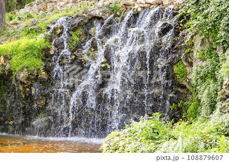 Waterfall. Waterfall in a park in Madrid. Artificial Mountain of Buen Retiro, called Mountain of the Cats. Dark stone background surrounded by green vegetation and plants and leaves. Autumn colors. Waterfall. Waterfall in a park in Madrid. Artificial Mountain of Buen Retiro, called Mountain of the Cats. Dark stone background surrounded by green vegetation and plants and leaves. Autumn colors. 108879607