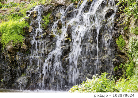 Waterfall. Waterfall in a park in Madrid. Artificial Mountain of Buen Retiro, called Mountain of the Cats. Dark stone background surrounded by green vegetation and plants and leaves. Autumn colors. 108879624