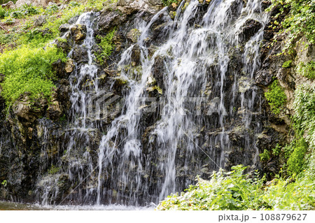 Waterfall. Waterfall in a park in Madrid. Artificial Mountain of Buen Retiro, called Mountain of the Cats. Dark stone background surrounded by green vegetation and plants and leaves. Autumn colors. Waterfall. Waterfall in a park in Madrid. Artificial Mountain of Buen Retiro, called Mountain of the Cats. Dark stone background surrounded by green vegetation and plants and leaves. Autumn colors. 108879627