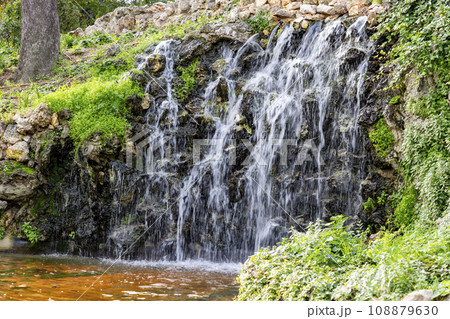 Waterfall. Waterfall in a park in Madrid. Artificial Mountain of Buen Retiro, called Mountain of the Cats. Dark stone background surrounded by green vegetation and plants and leaves. Autumn colors. Waterfall. Waterfall in a park in Madrid. Artificial Mountain of Buen Retiro, called Mountain of the Cats. Dark stone background surrounded by green vegetation and plants and leaves. Autumn colors. 108879630